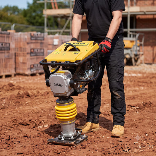 A person wearing work gloves operates a yellow and silver RM-80 plate compactor
