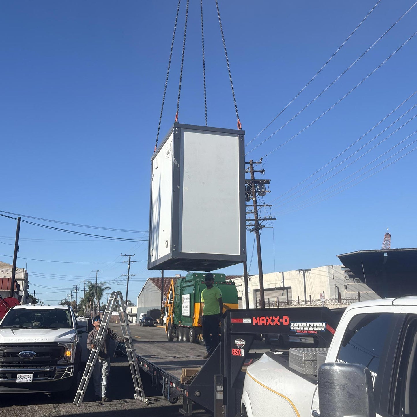 A portable restroom is being lifted by a crane onto a flatbed trailer