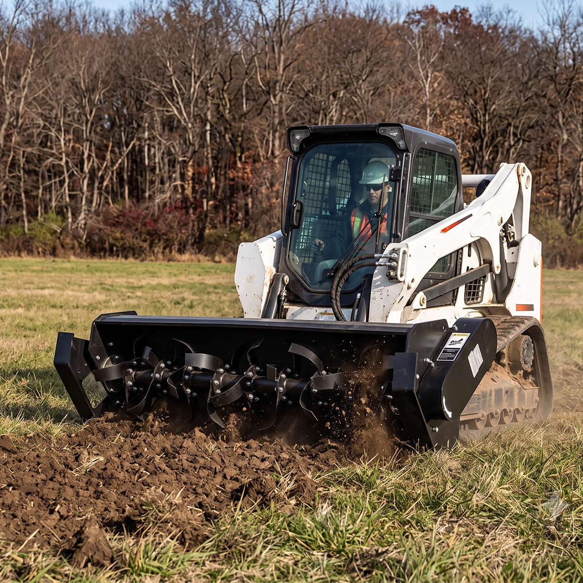 A white Bobcat skid steer loader with a rotary tiller attachment is operating in a grassy field, turning dark brown soil. 