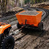 Orange dump trailer attached to a four-wheeler on a muddy trail with gravel load